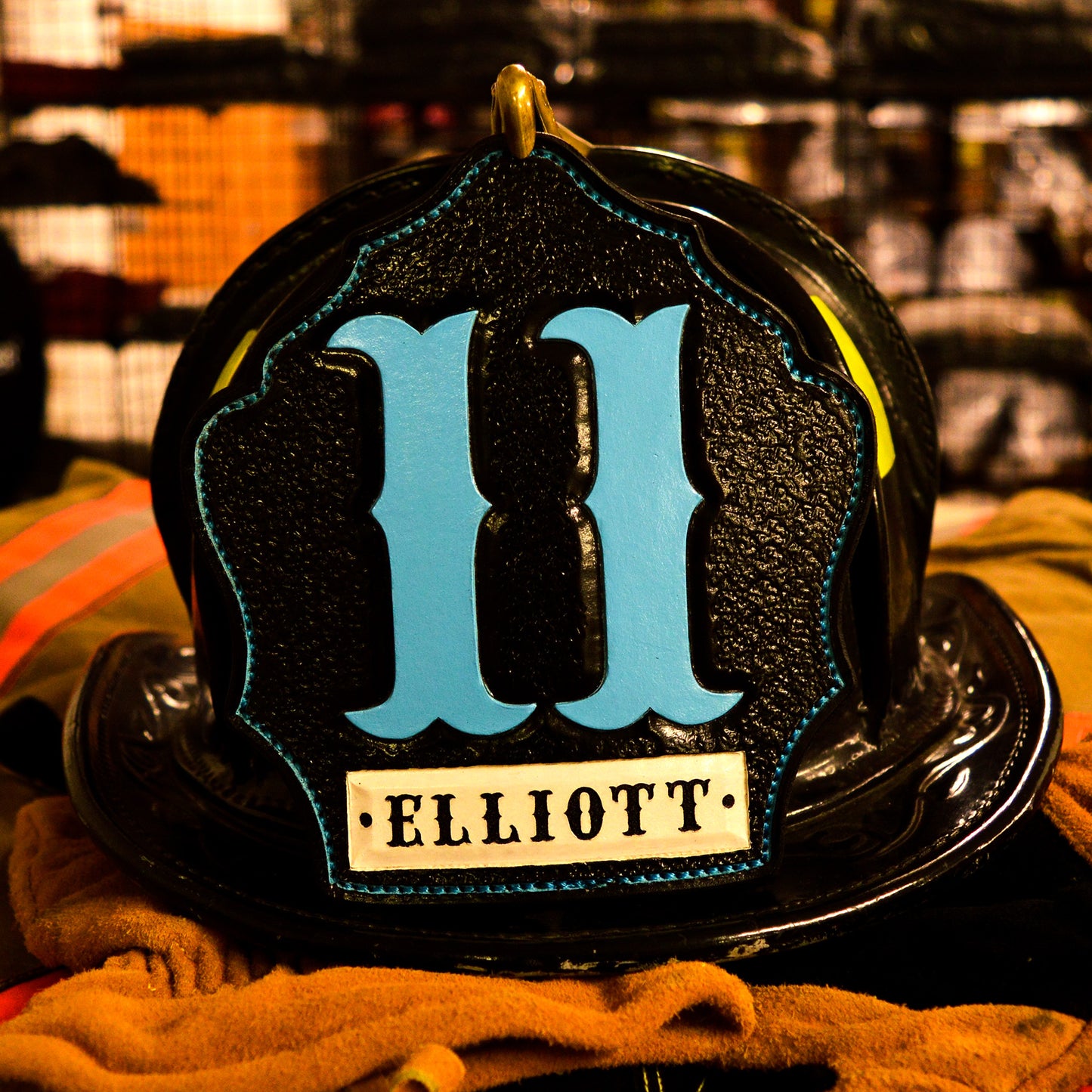 Black leather firefighter helmet shield with light blue numerals “11,” teal outline accents, and a white nameplate reading ELLIOTT—mounted on a helmet