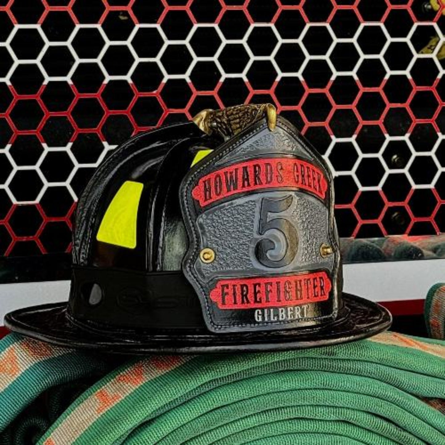 Custom leather firefighter helmet shield with raised numeral and red top/bottom banners on a textured gray panel—mounted on a black fire helmet with hose in the background.