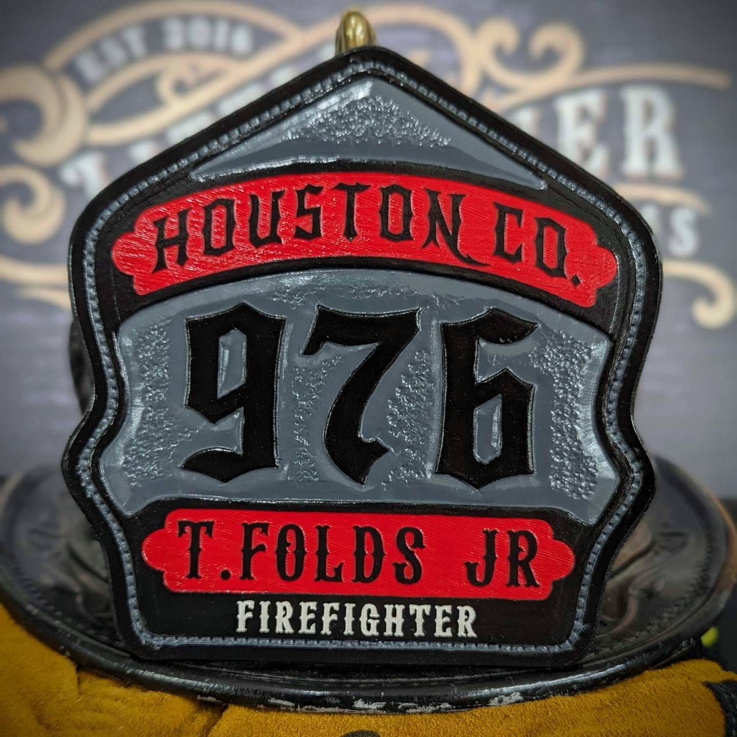 Custom leather firefighter helmet shield with textured gray center panel, red top and bottom banners, and raised black numerals—hand-painted and mounted on a helmet.