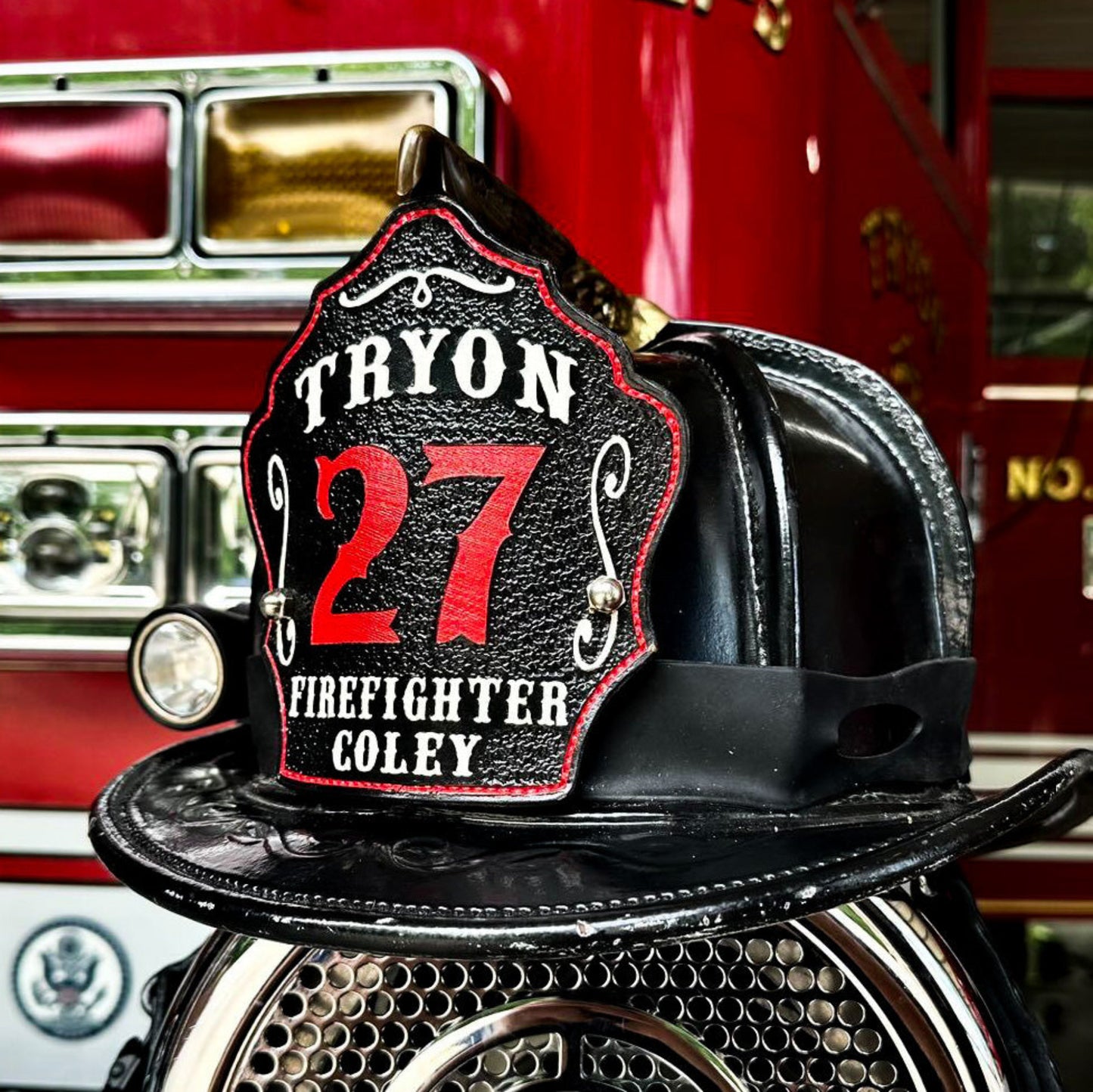 Custom leather firefighter helmet shield — Tryon Fire Department — black shield with red “27” and white “FIREFIGHTER COLEY,” hand-painted and mounted on a traditional black leather helmet in front of a red fire engine by Little River Customs.