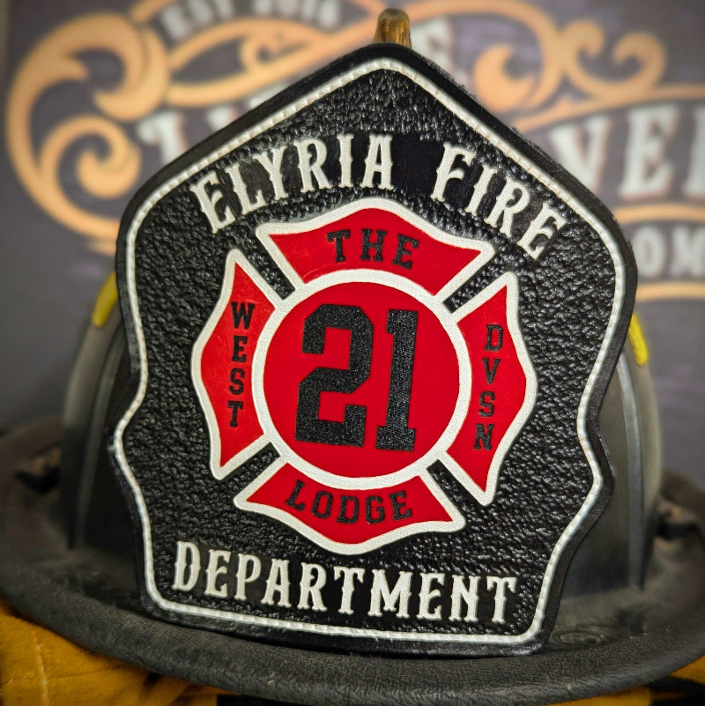 Black leather firefighter helmet shield featuring a red Maltese cross with bold number 21, Elyria Fire Department, The West Lodge Division text accents.