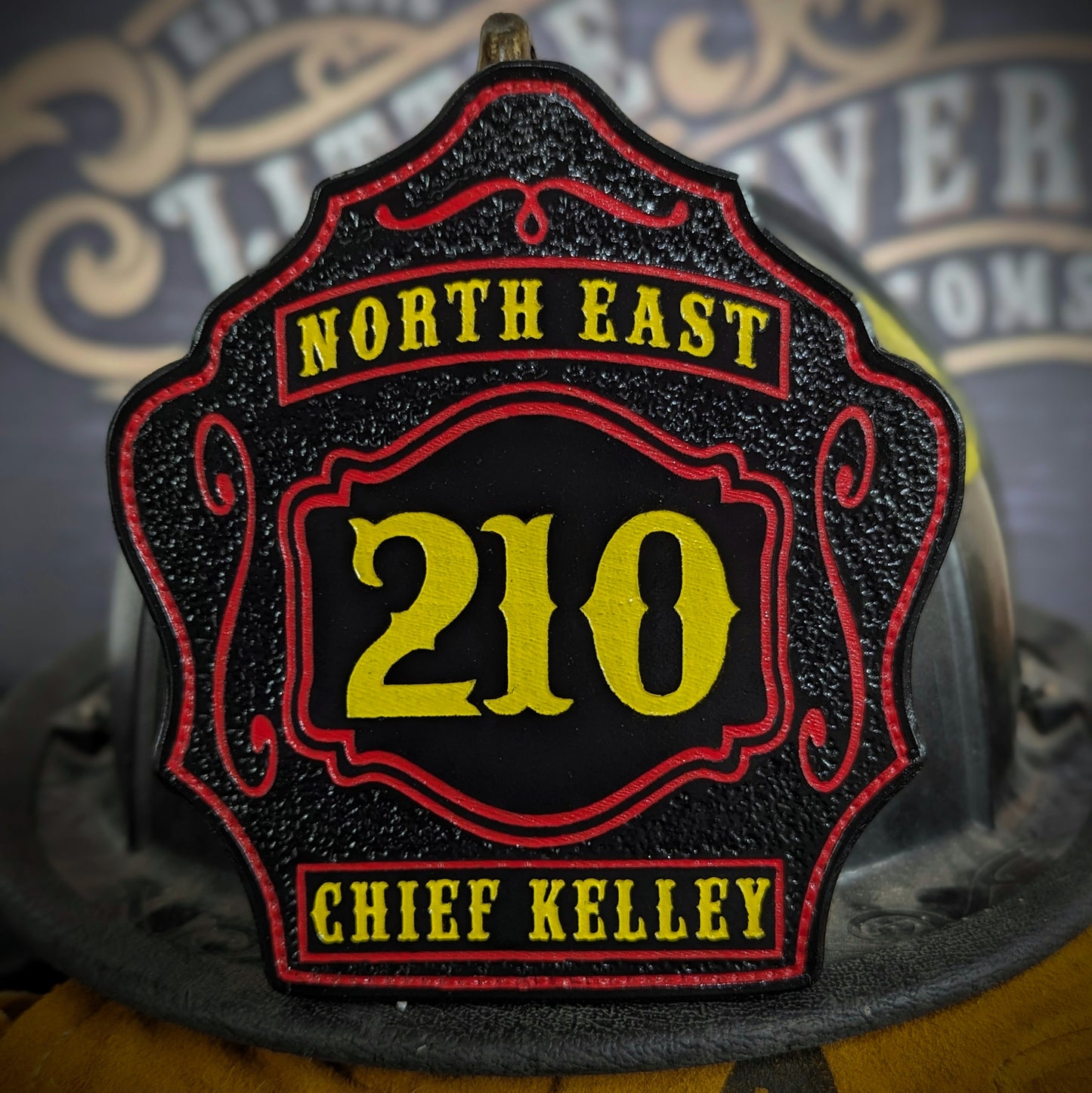 Black leather firefighter helmet shield with bold yellow number 210, red trim and scrolls, North East text at top, and Chief Kelley nameplate at bottom.