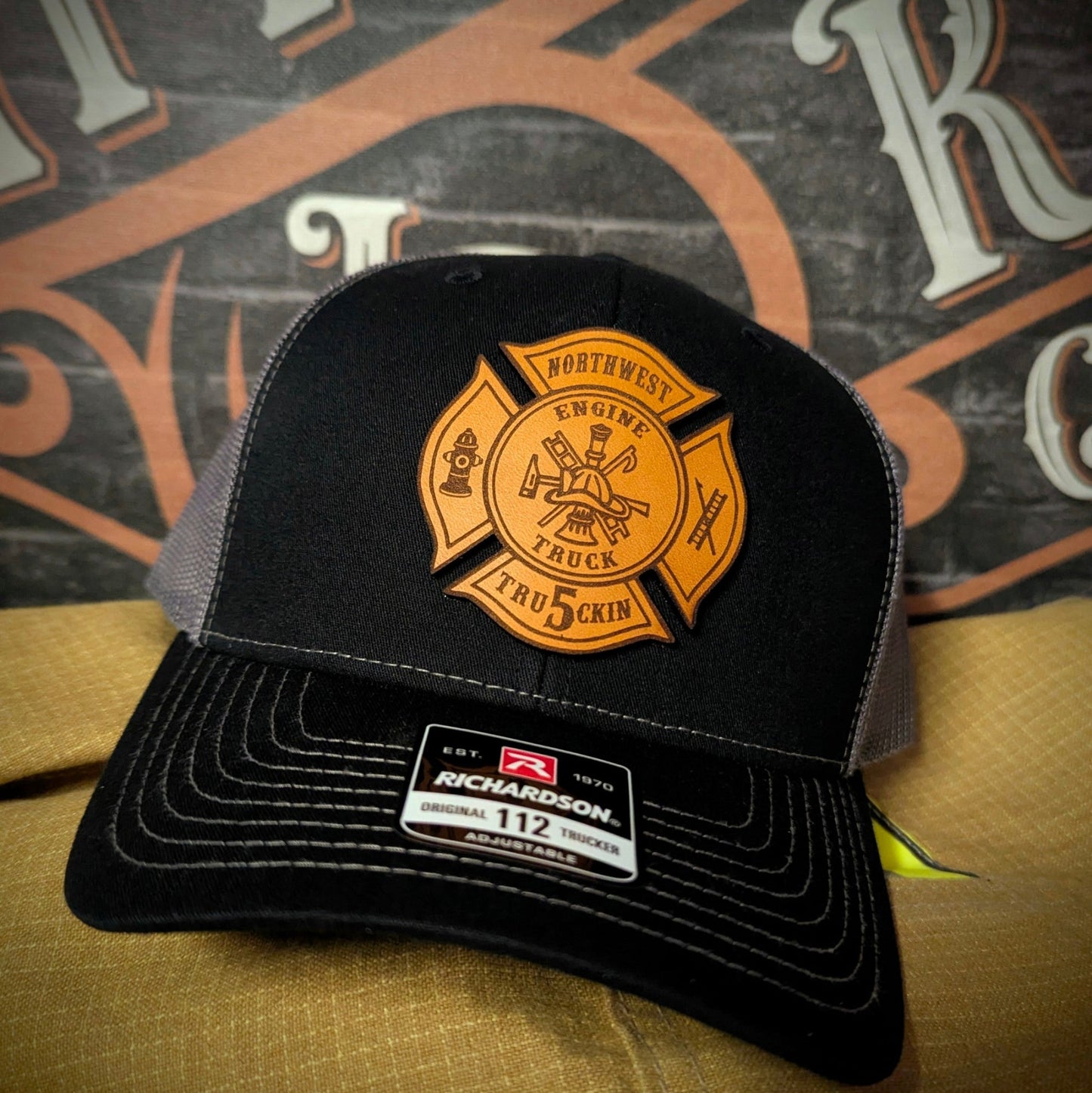 Black and gray Richardson firefighter hat with custom Maltese leather patch design, rugged headwear for fire service crews.
