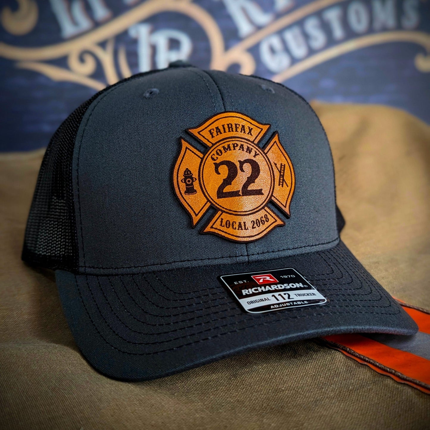 Charcoal Richardson firefighter hat with custom Maltese leather patch design, rugged mesh back style for fire service crews.