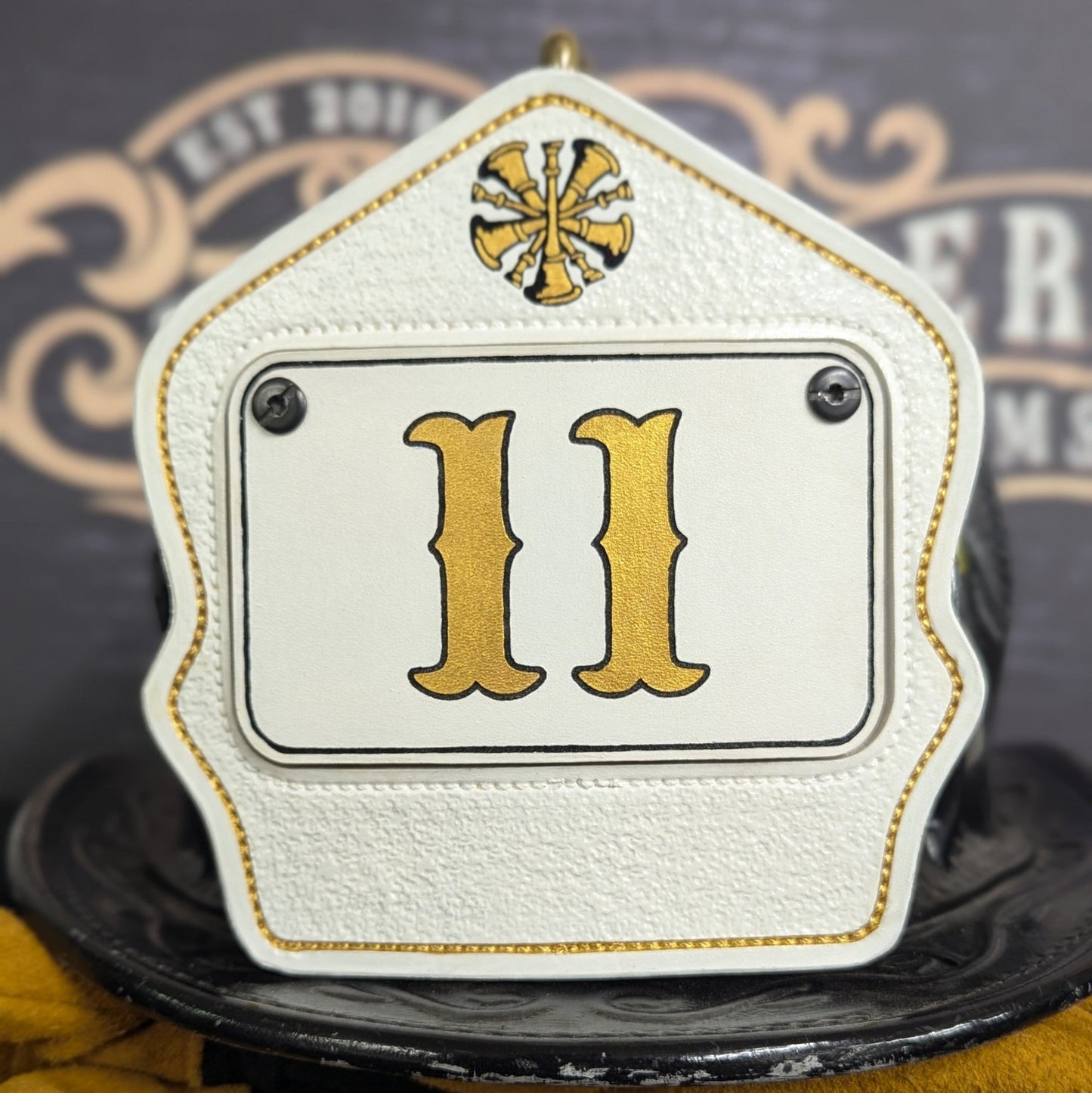 White leather firefighter helmet shield with gold numerals on a white inset panel, bugle crest at the top, and gold trim—mounted on a helmet.