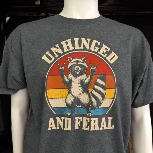 Dark gray t-shirt with retro-style raccoon graphic and “Unhinged and Feral” text – funny feral vibes shirt with rock-on hand gesture