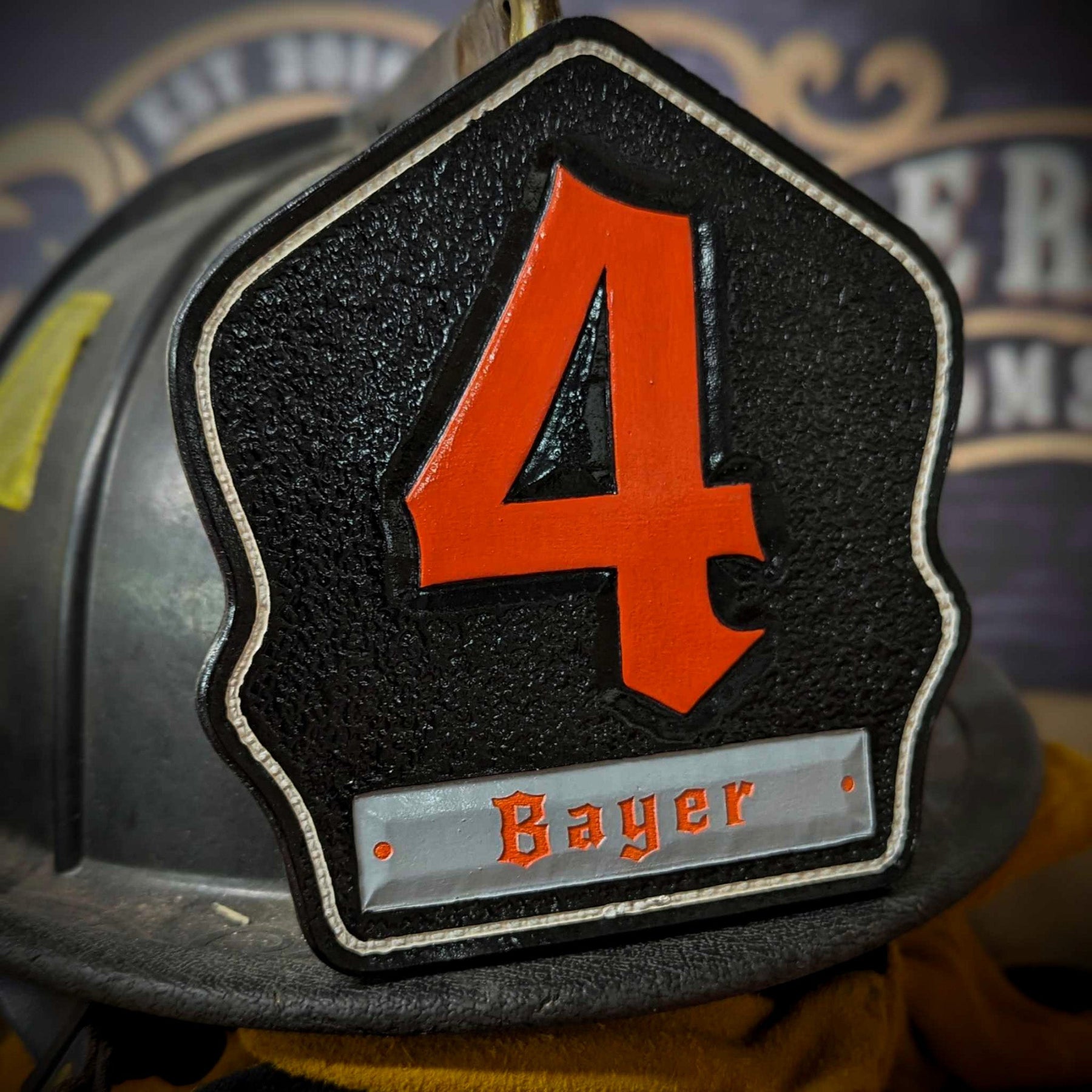 Black leather firefighter helmet shield with large red numeral “4” and a gray nameplate with orange lettering—mounted on a helmet.