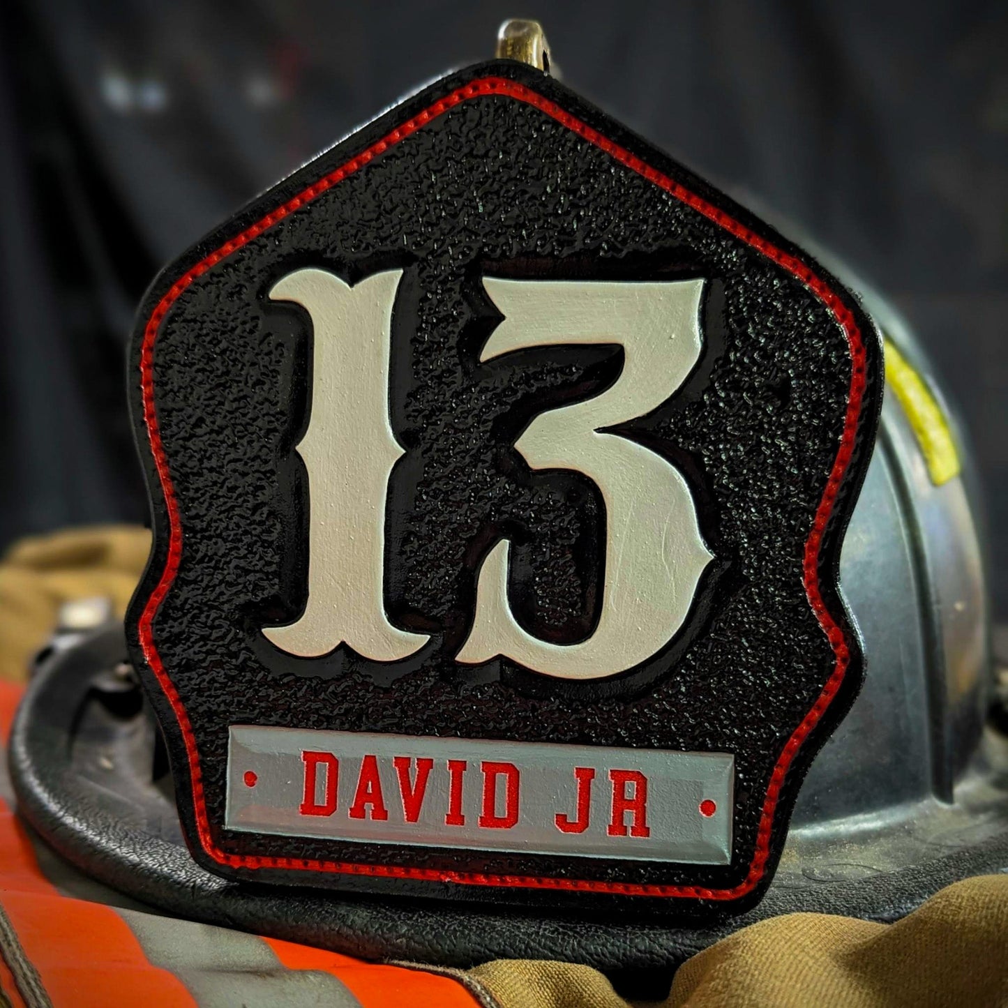 Black leather firefighter helmet shield with large white numerals “13,” red border outline, and gray nameplate reading DAVID JR—mounted on a helmet.