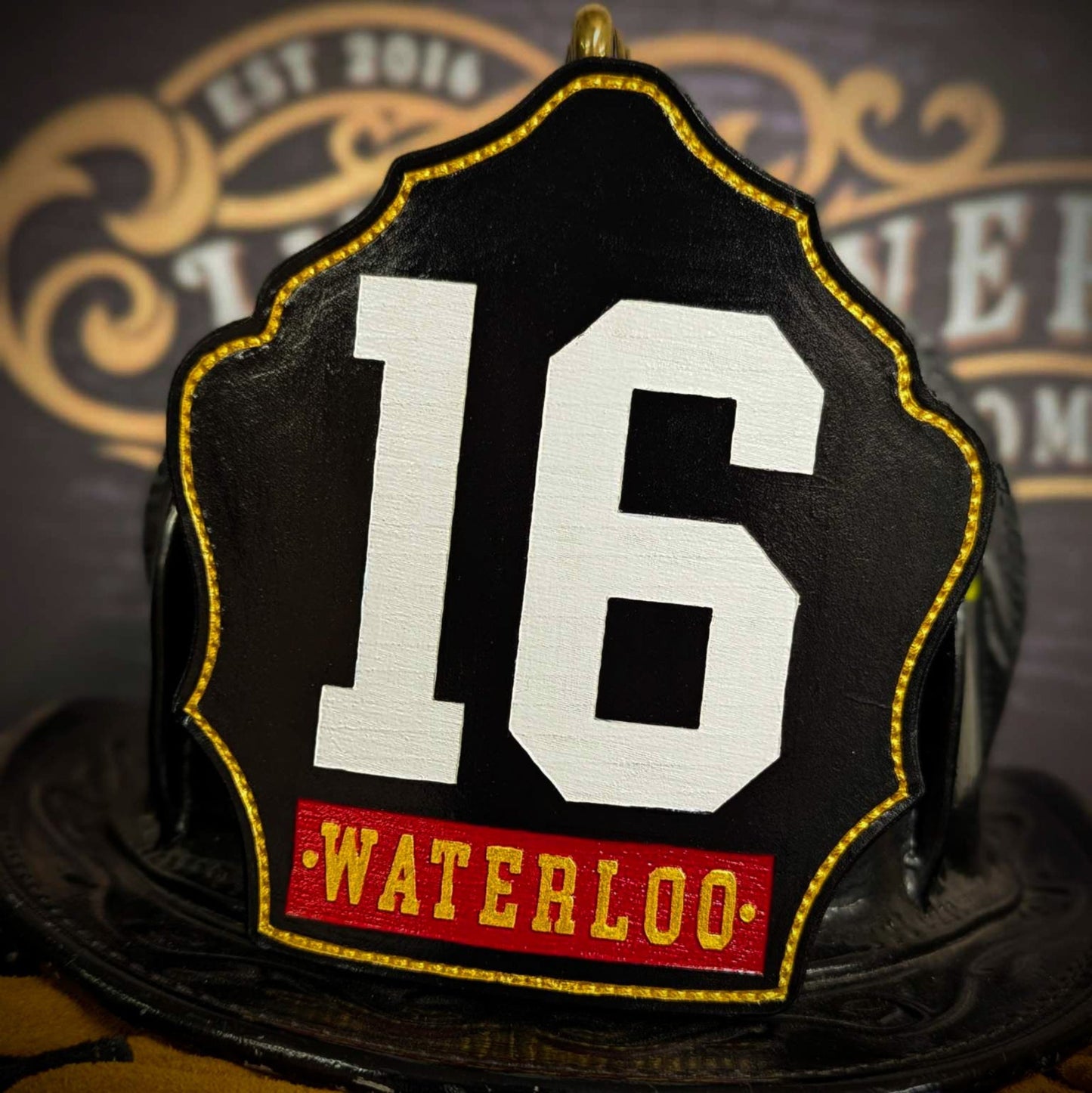 Black leather firefighter helmet shield with large white numerals “16,” gold stitched edge, and red nameplate reading WATERLOO—mounted on a helmet.