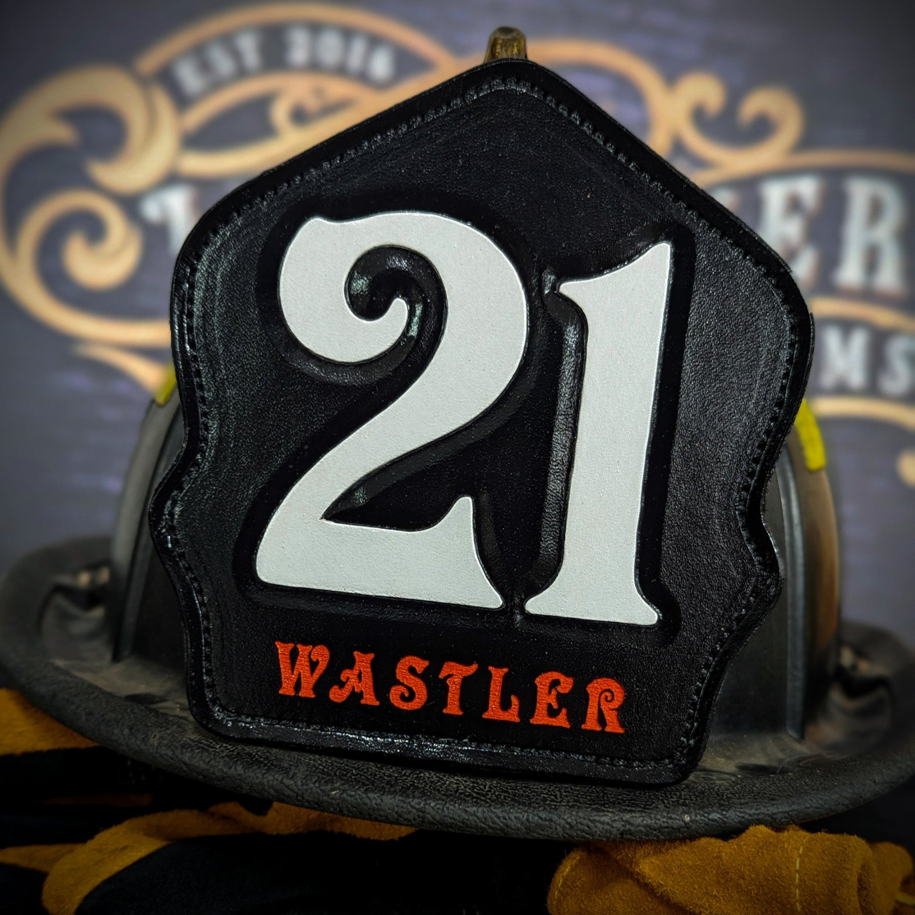 Black leather firefighter helmet shield with large white numerals “21” and orange name lettering—mounted on a helmet.