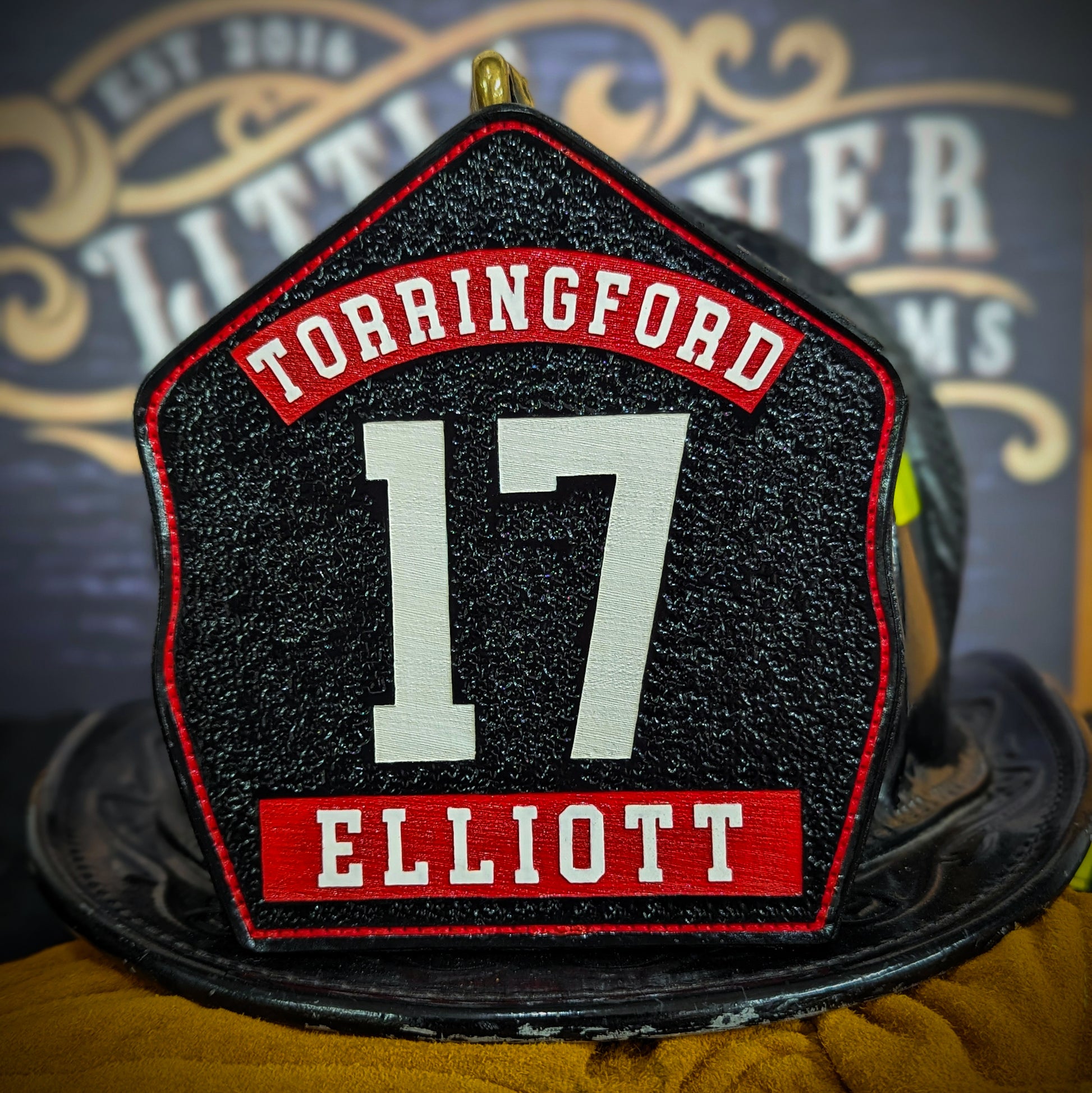 Black leather firefighter helmet shield with large white number 17, red Torringford banner at top, and red nameplate with Elliott at bottom.