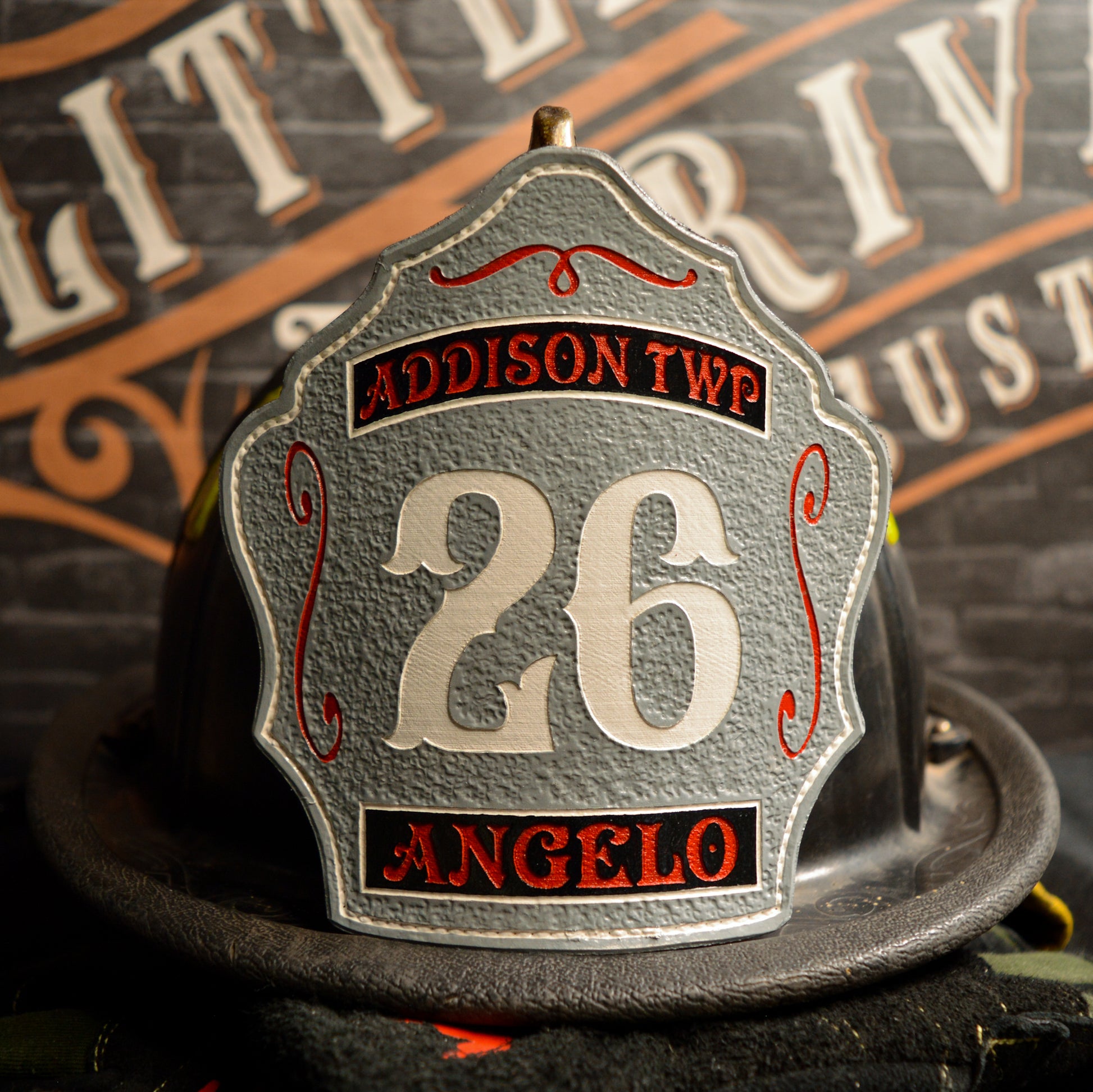 Gray leather firefighter helmet shield with large white number 26, top banner Addison TWP in red on black, bottom banner Angelo in red, with red scroll accents.