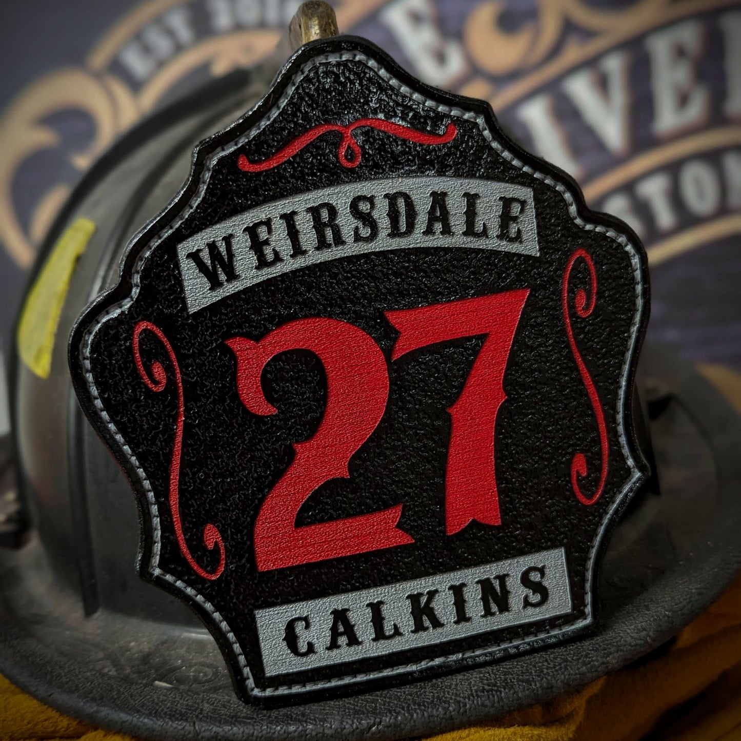 Custom black leather firefighter helmet shield with number 27 in red, gray banners, and decorative scroll accents