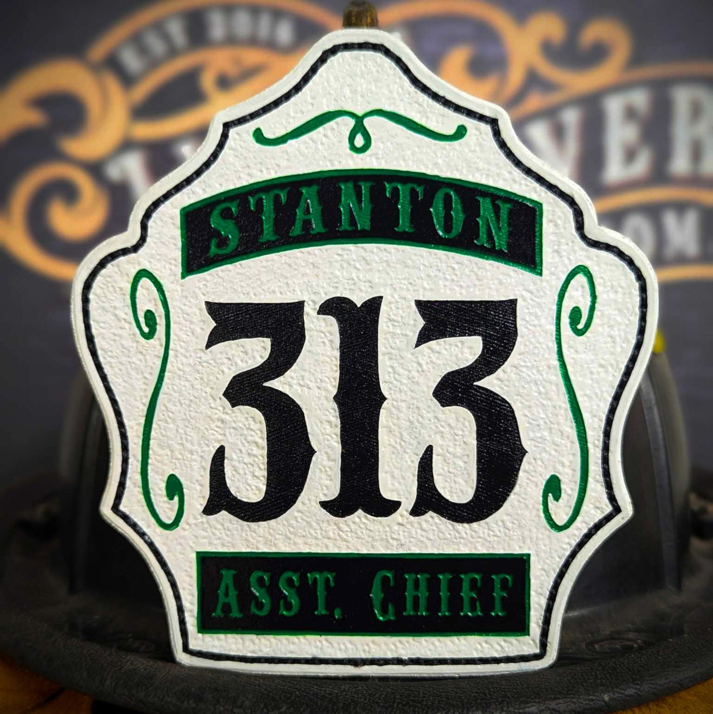 White leather firefighter helmet shield with bold black number 313, green banners reading Stanton and Asst. Chief, and green scroll accents.