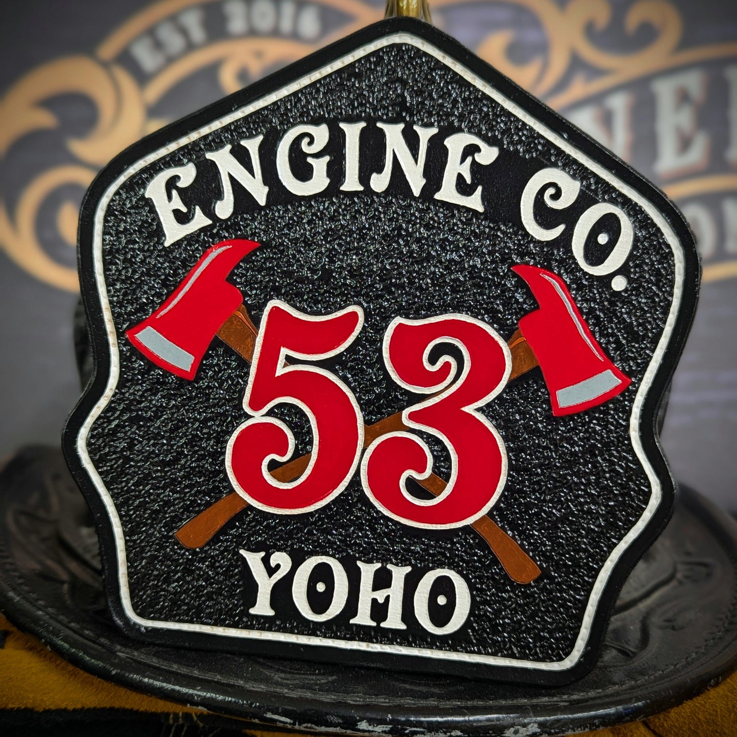 Black leather firefighter helmet shield with crossed red axes and bold red number 53, custom fire department gear.