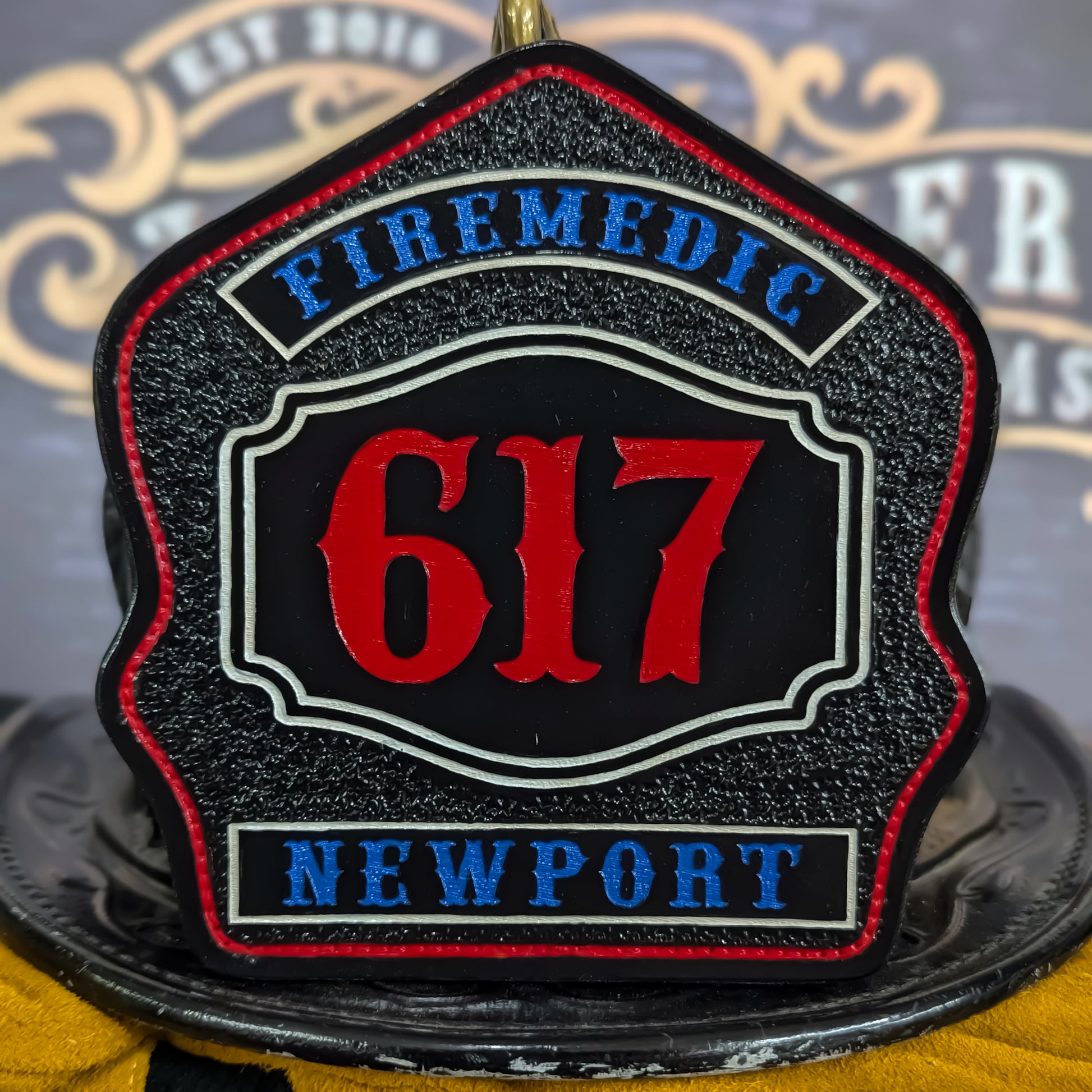 Custom black leather firefighter helmet shield with bold red number 617, blue text reading Firemedic and Newport, accented with white borders and mounted on a helmet.