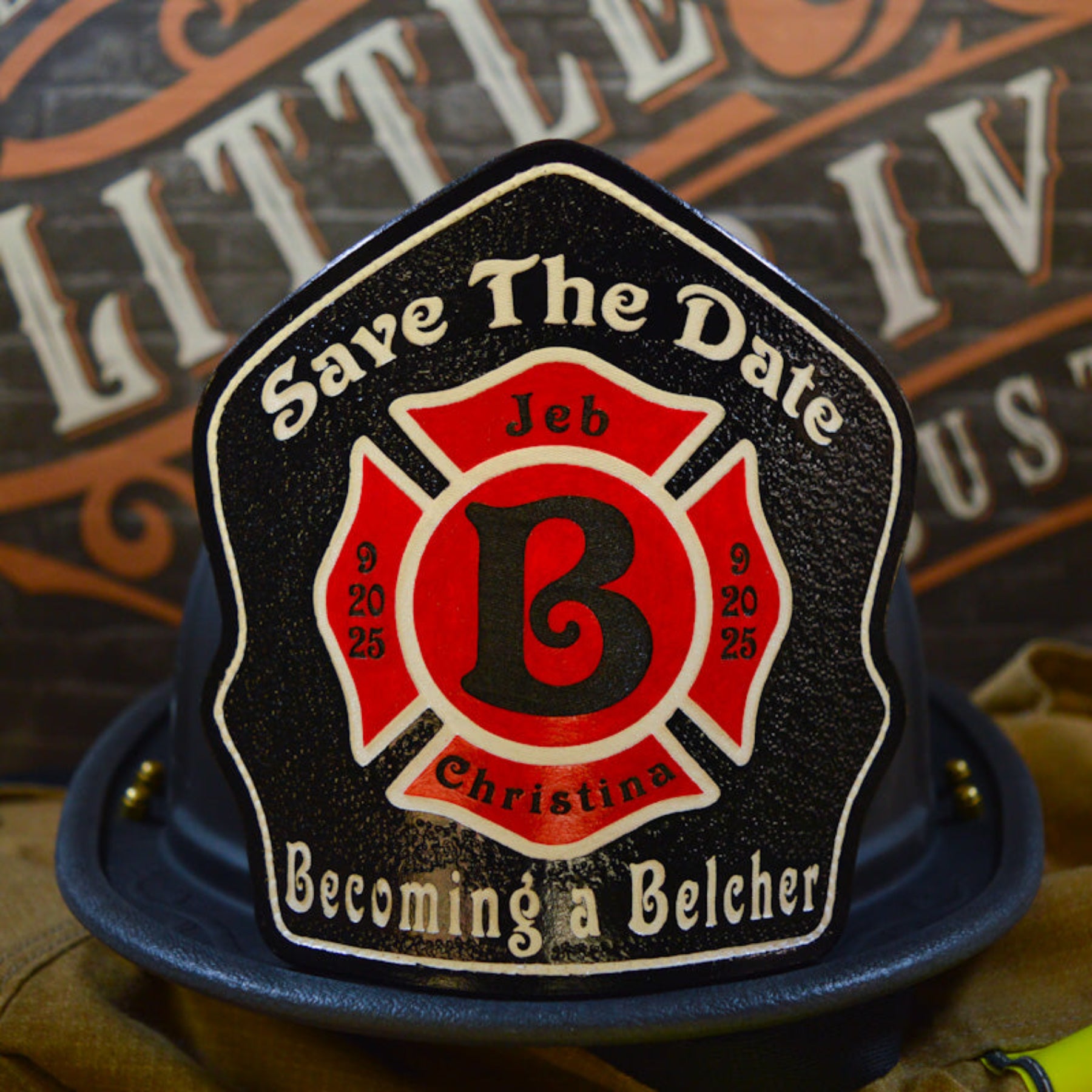 Custom firefighter helmet shield with a black background, red Maltese cross, bold letter B, and personalized wedding details for Jeb and Christina with "Save The Date" and "Becoming a Belcher" text.