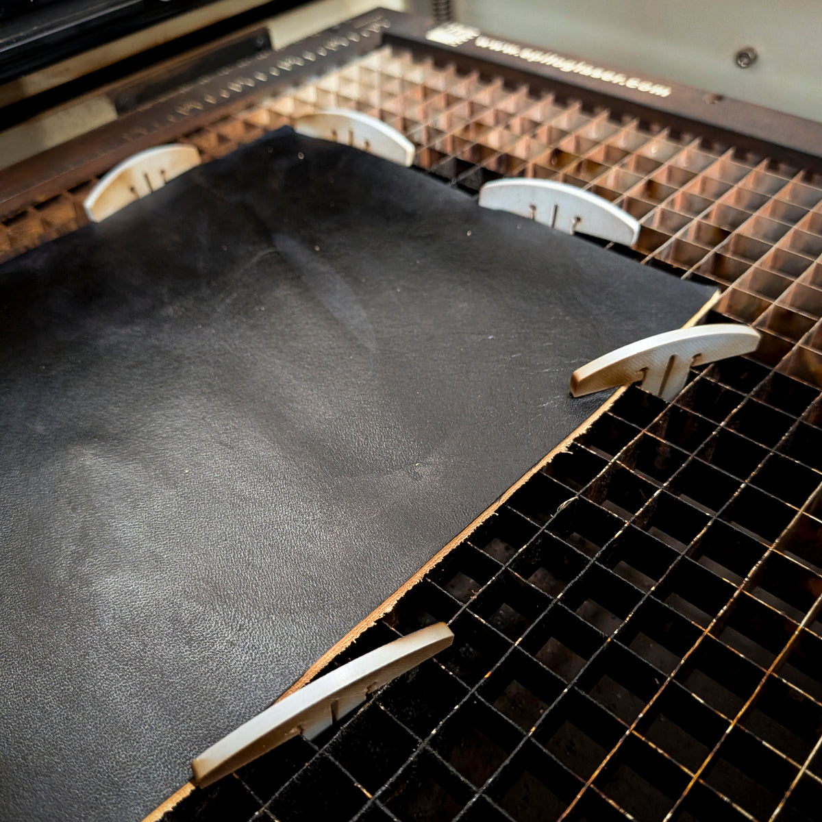 Laser bed dogs holding a leather sheet flat on a honeycomb cutting bed.