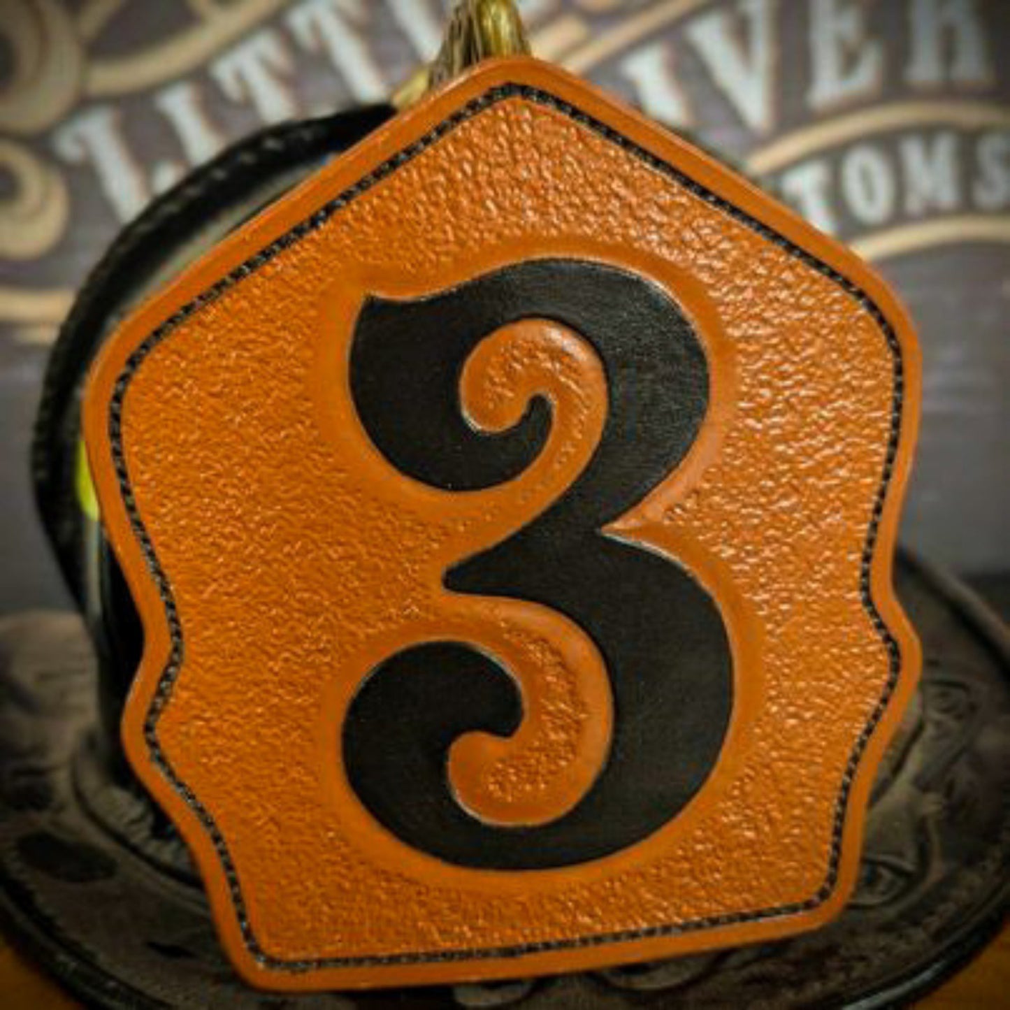 Tan leather firefighter helmet shield with large black numeral “3” and black stitching—textured face, mounted on a helmet.