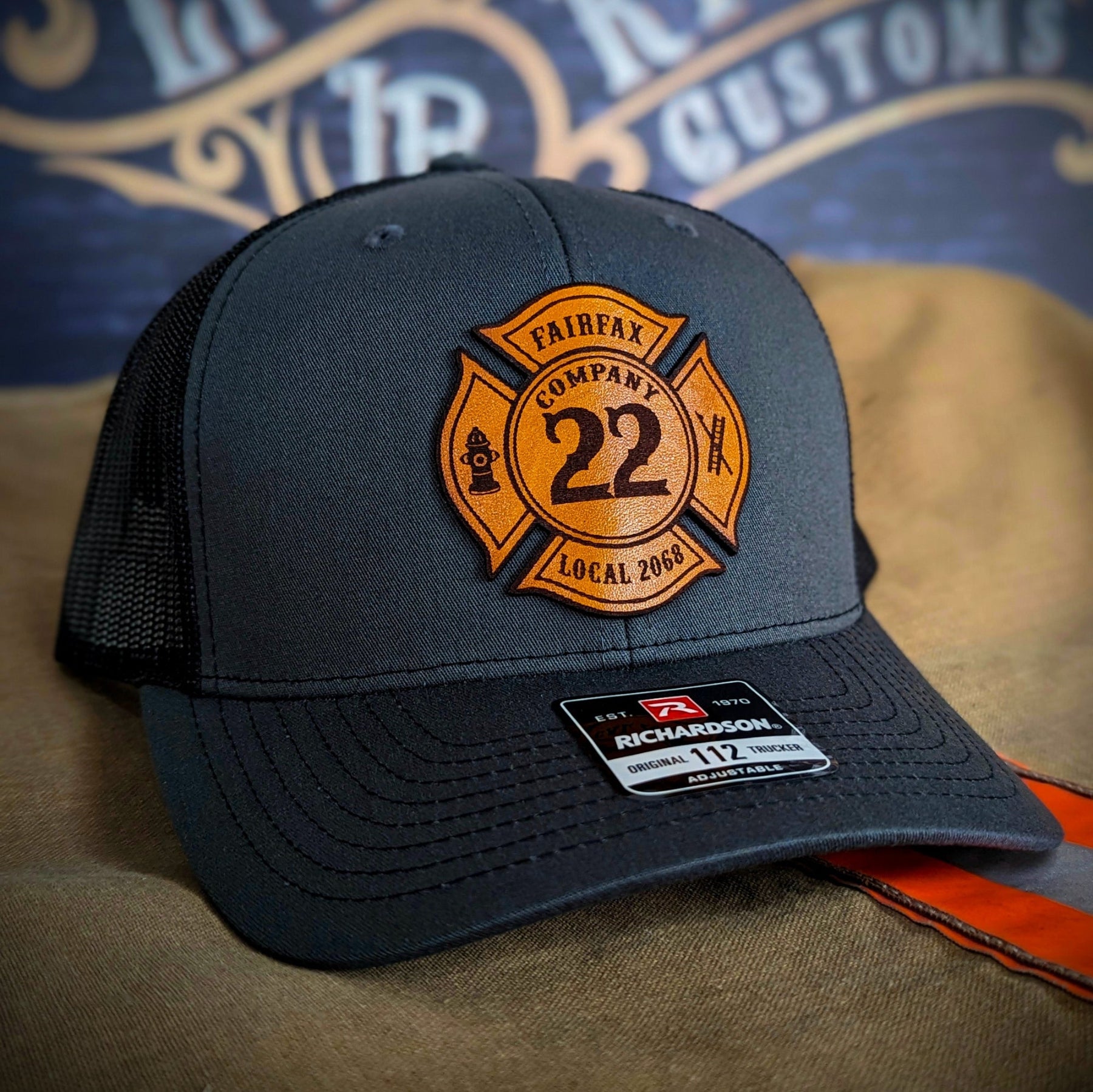 Charcoal Richardson firefighter hat with custom Maltese leather patch design, rugged mesh back style for fire service crews.