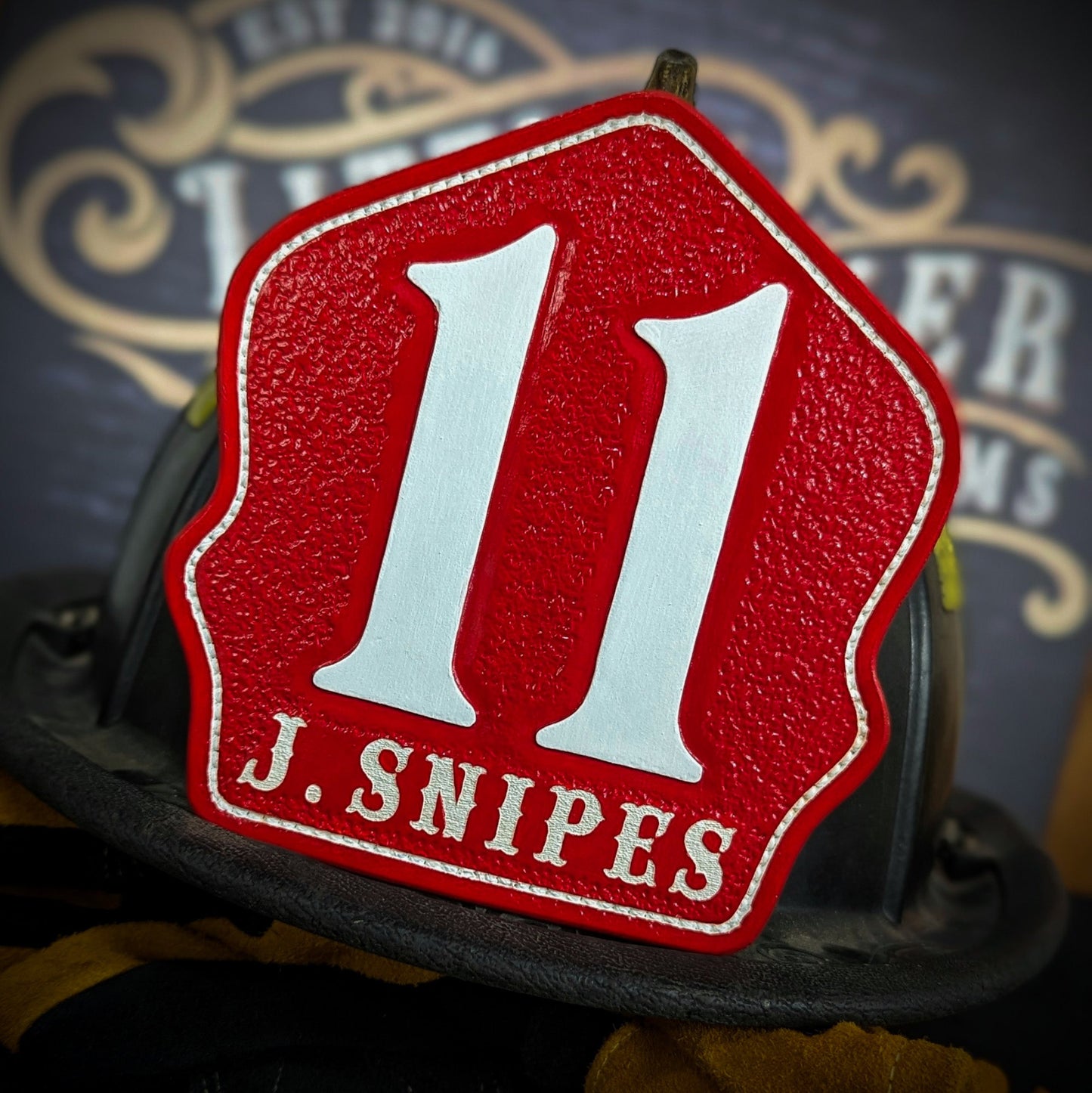 Red leather firefighter helmet shield with large white numerals “11” and white name lettering “J. SNIPES”—mounted on a helmet.
