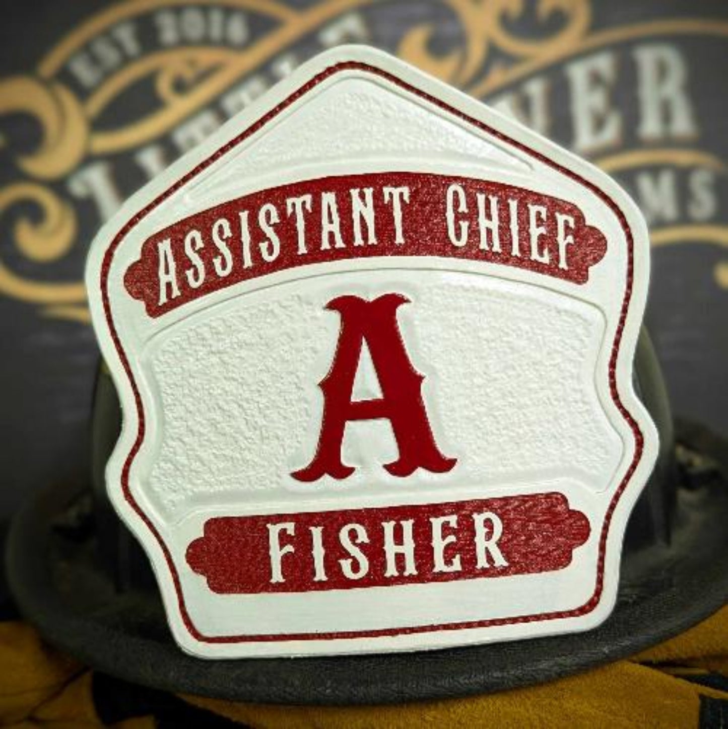 White chief-style leather firefighter helmet shield with red initial and banner accents—hand-painted and mounted on a helmet.