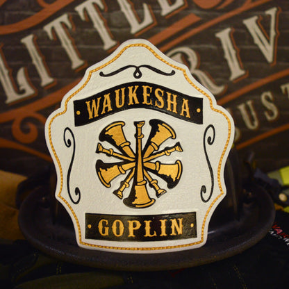 White leather firefighter helmet shield with crossed gold bugles, black title/name banners, and gold trim—hand-painted and mounted on a helmet.