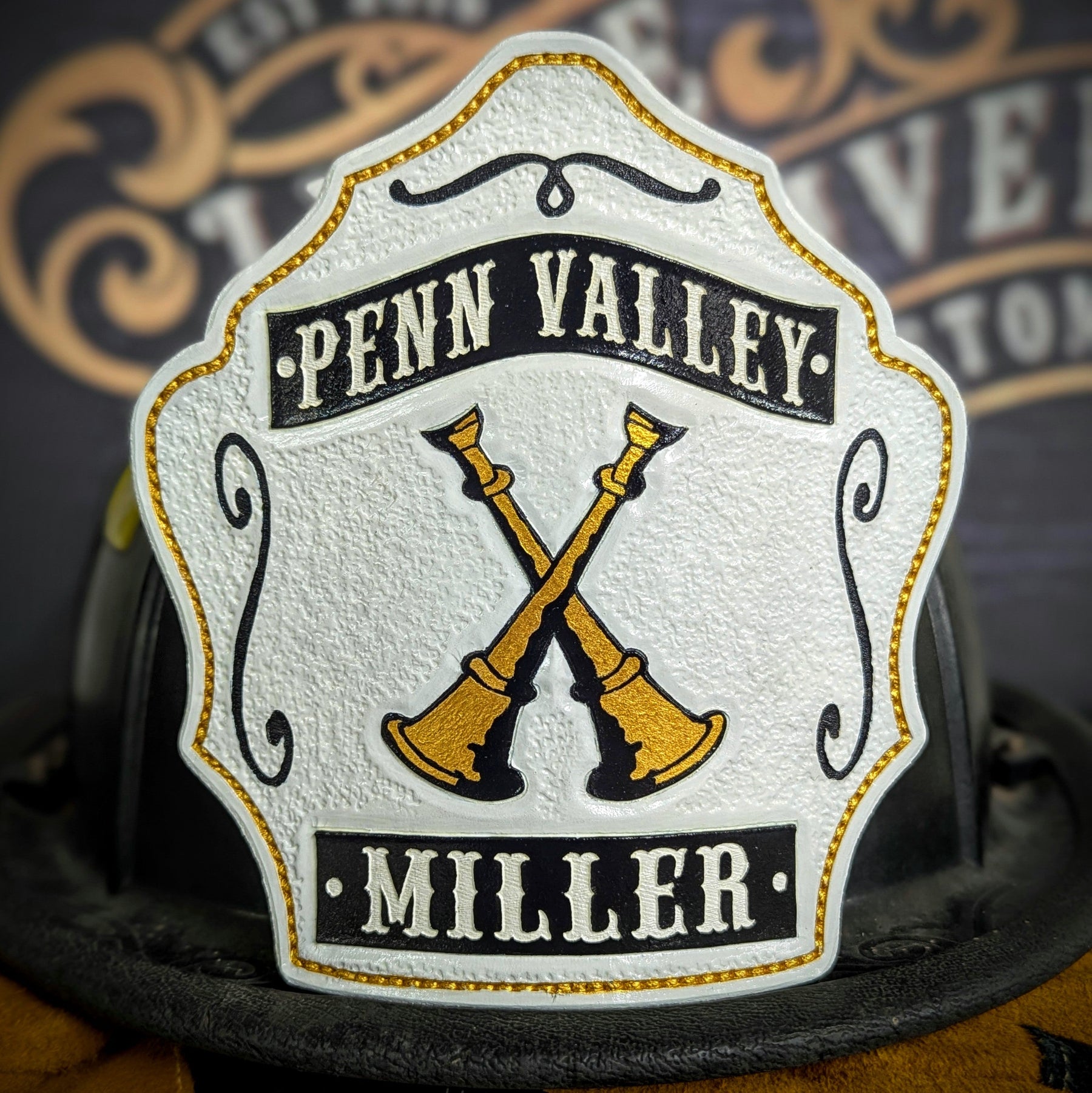White leather firefighter helmet shield with crossed gold bugles and black title/name banners—hand-painted on a textured face, mounted on a helmet.