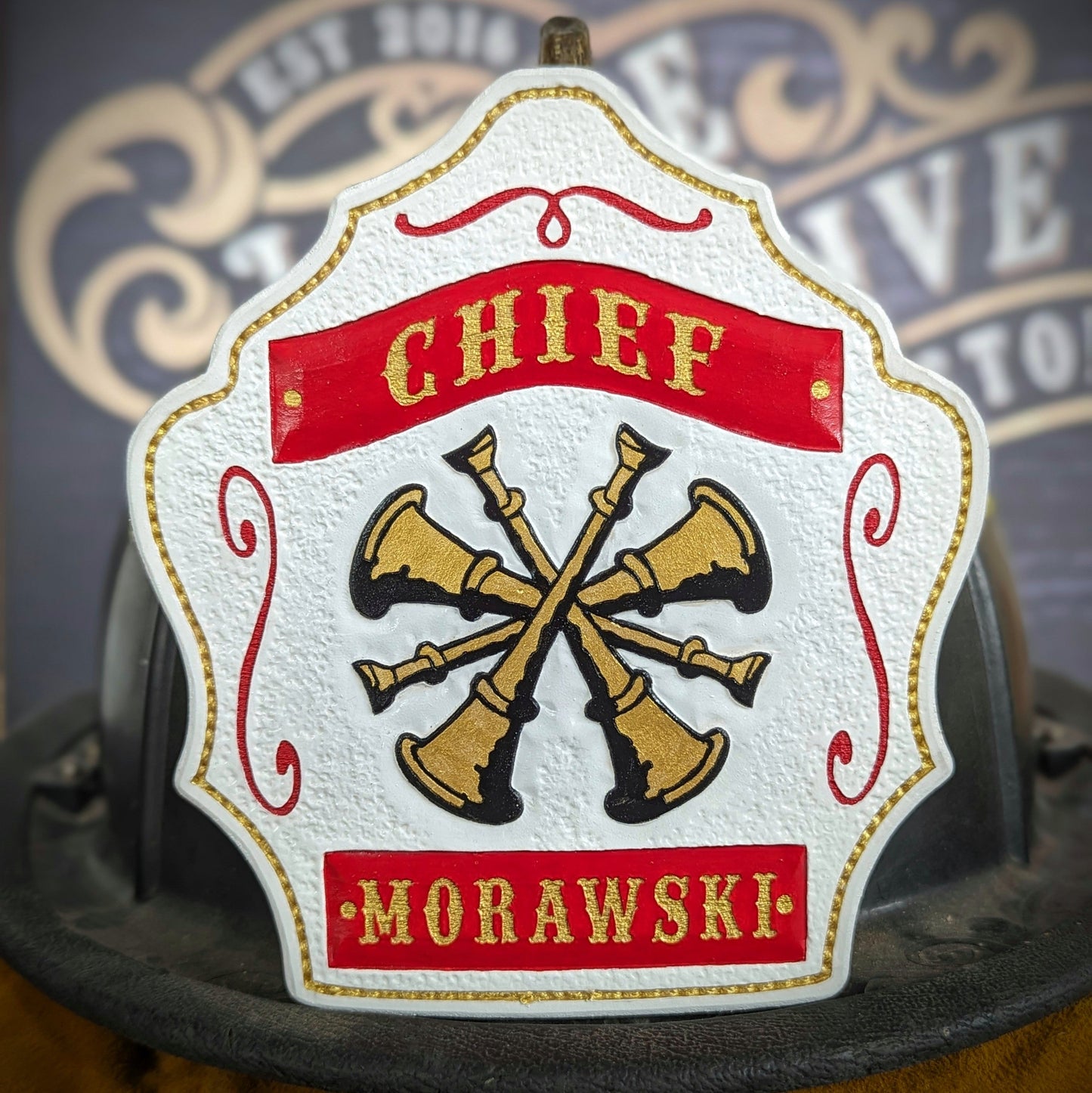 White leather firefighter helmet shield with crossed gold bugles and red banners—hand-painted and mounted on a helmet