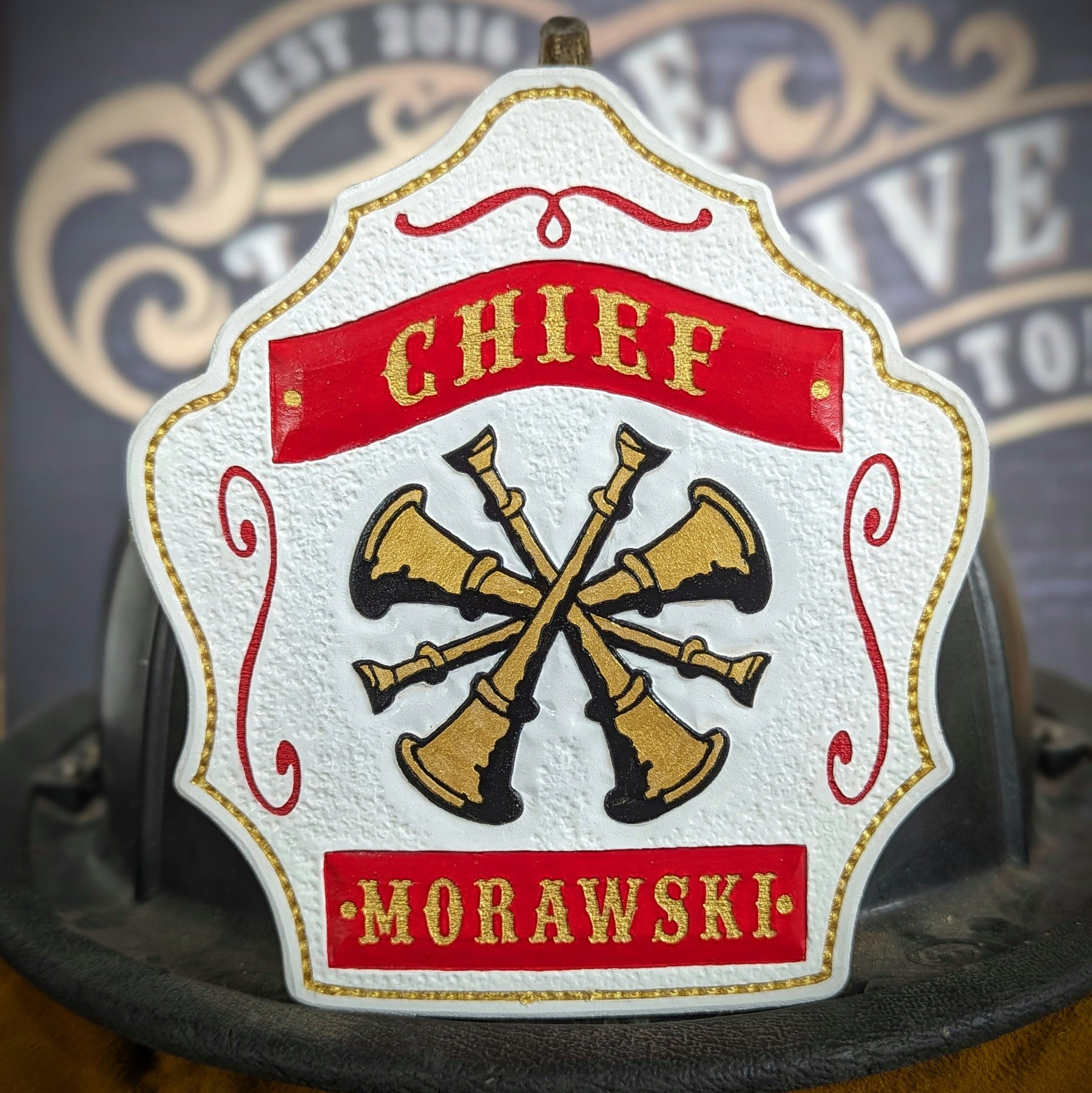White leather firefighter helmet shield with crossed gold bugles and red banners—hand-painted and mounted on a helmet