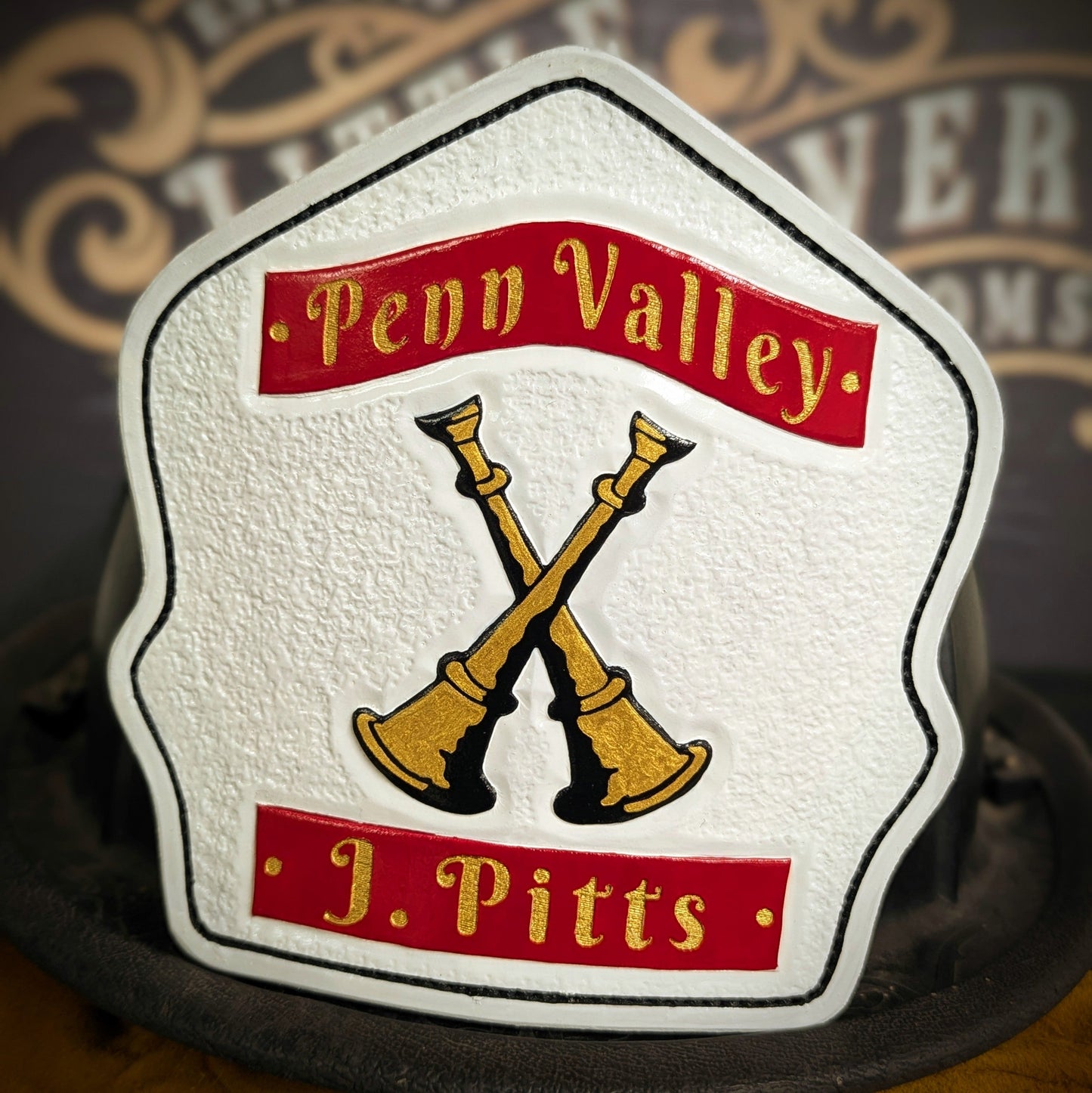 White leather firefighter helmet shield with crossed gold bugles and red title/name banners—hand-painted and mounted on a helmet.