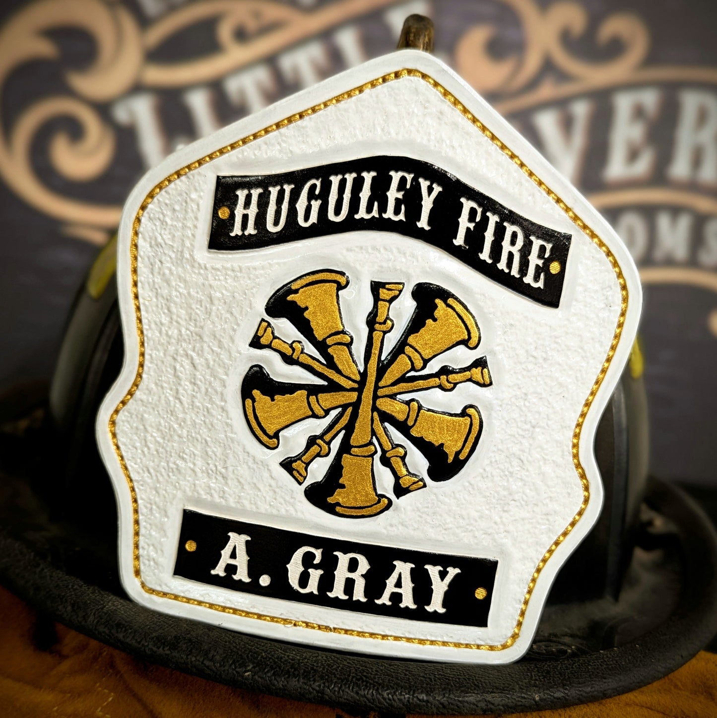White leather firefighter helmet shield with gold bugle emblem and black title/name banners—hand-painted and mounted on a helmet.