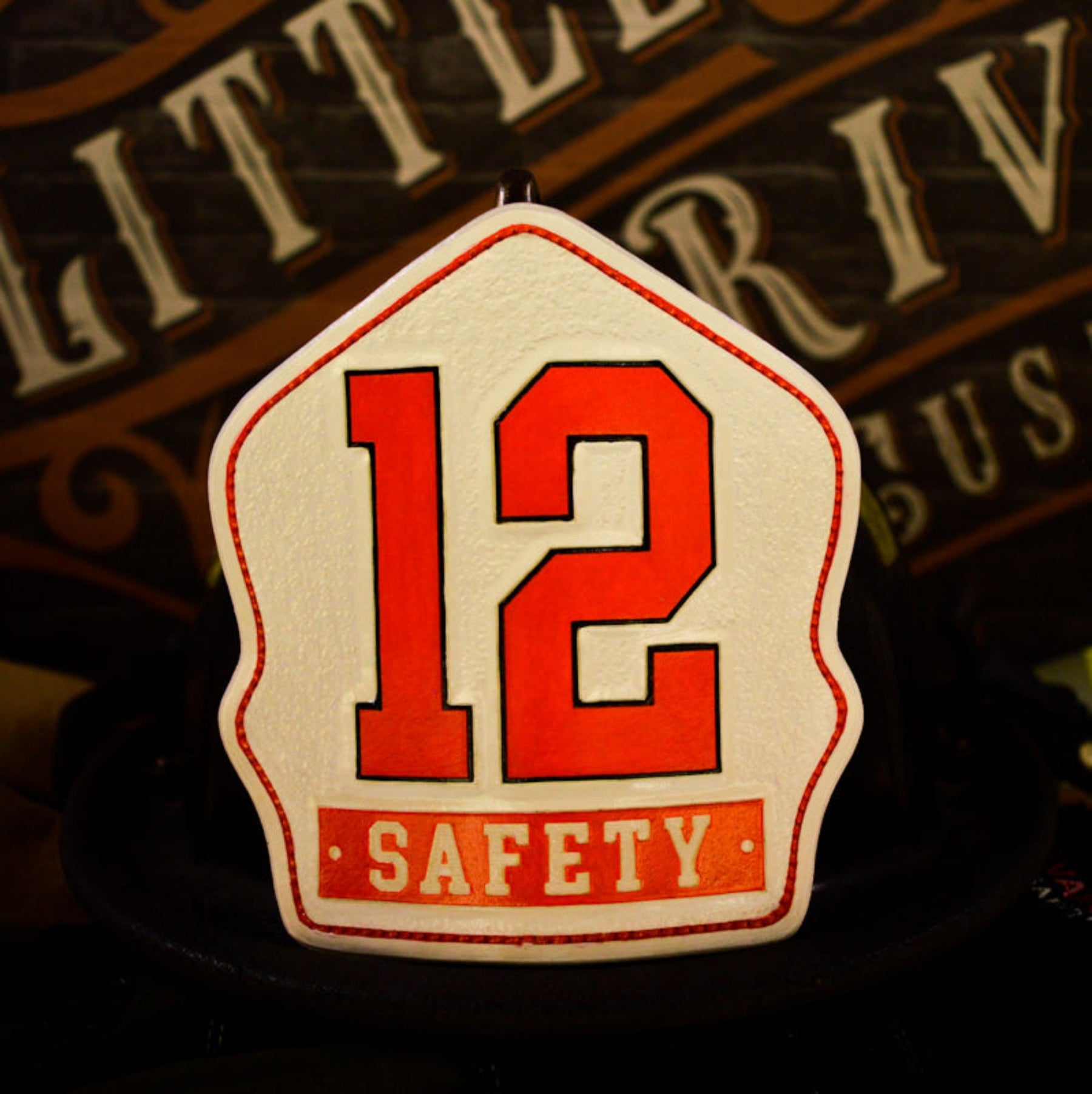 White leather firefighter helmet shield with bold red numerals “12” and a red “SAFETY” banner—hand-painted and mounted on a helmet.